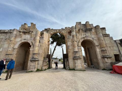       Large stone Roman gateway with twin arches framing visitors and a solitary tree in ancient ruins.
  