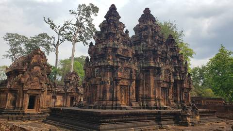       Intricately carved red-sandstone towers of Banteay Srei temple amid greenery under a cloudy sky
  