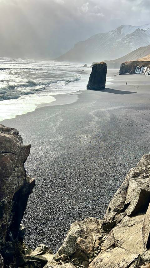       Close-up view of a dark volcanic sand beach with gentle surf and foam lines.
  