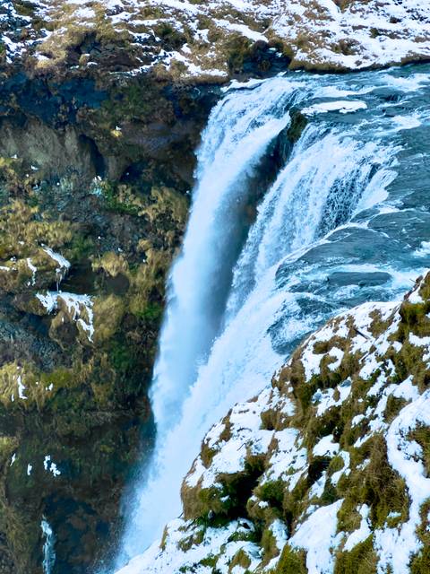       Side view of a tall waterfall plunging down mossy cliffs partially dotted with snow.
  