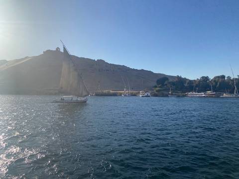       Traditional sailboat glides on the Nile with desert hills and ruins in the background.
  