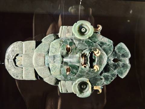       Intricately crafted jade funerary mask from ancient Maya civilization displayed in a case.
  