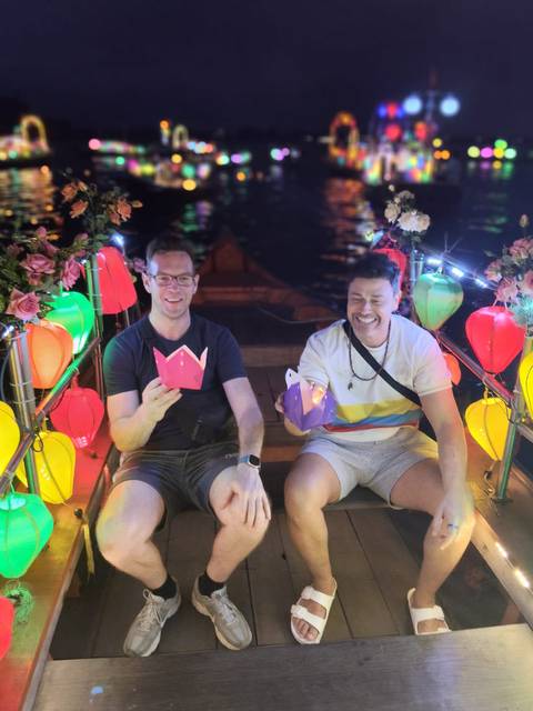       Two travelers on a lantern-lit boat in Hoi An holding paper flower lanterns at night.
  