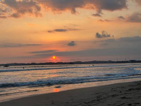       An orange sun sets over gentle waves and a distant shoreline, painting the sky in pastel hues.
  