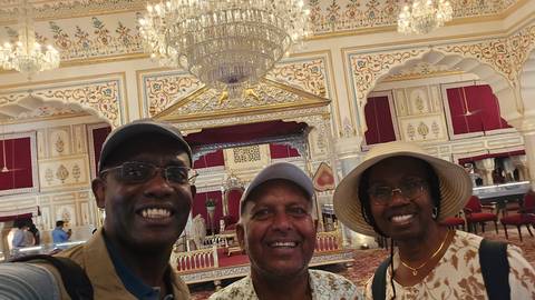       Three smiling travellers posing inside an ornately decorated palace hall with chandeliers.
  