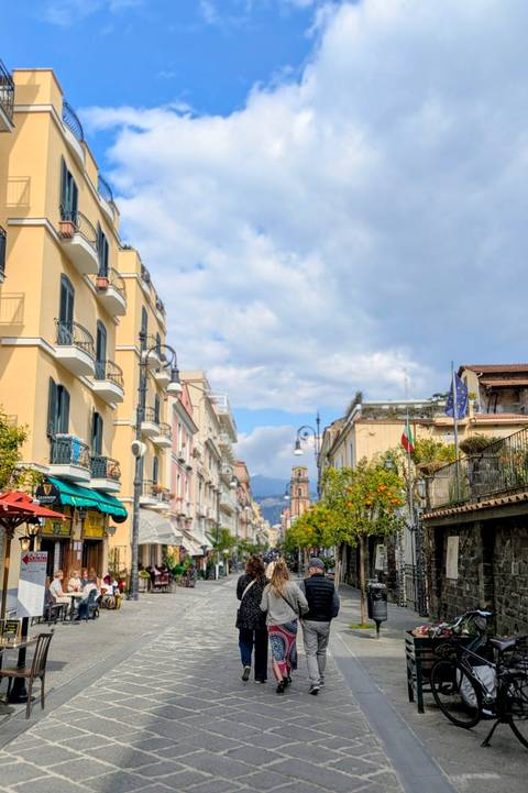       Charming pastel buildings line a pedestrian street in Sorrento with citrus trees and distant mountains.
  