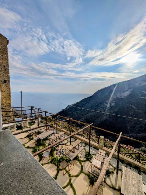       Terrace with wooden railings overlooking rugged coastal mountains and the shimmering Tyrrhenian Sea.
  