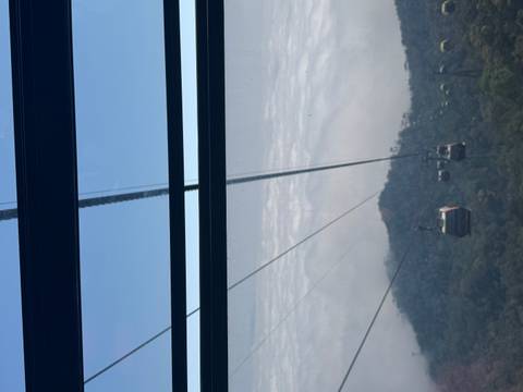       View through cable-car window of gondolas gliding above cloud layer and forested mountain slopes.
  