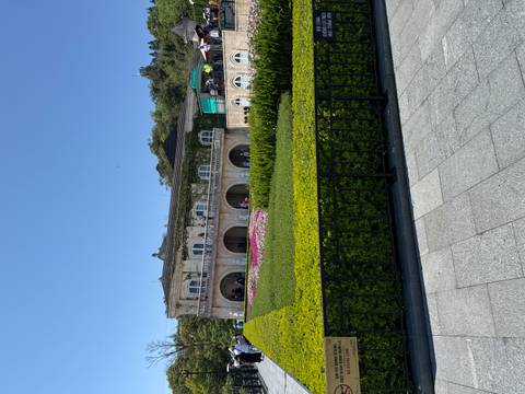       Neo-classical building framed by manicured hedges and colourful flower beds within a hilltop park.
  