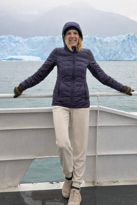       Woman in a puffer jacket poses with arms outstretched on the deck of a boat with cold grey water behind.
  