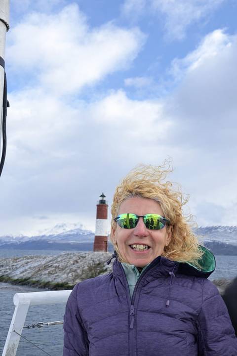      Close-up of a smiling traveler wearing mirrored sunglasses with the Les Éclaireurs lighthouse blurred in the background.
  