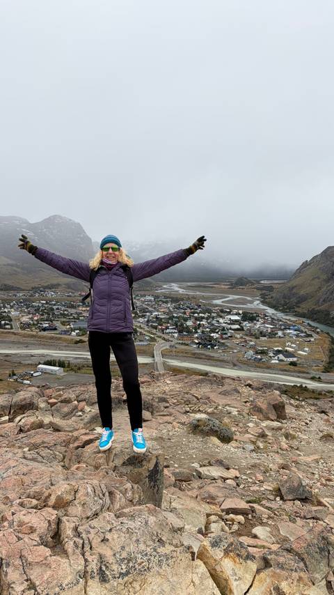       Excited hiker raises arms high above a Patagonian town stretching along a river valley framed by peaks.
  