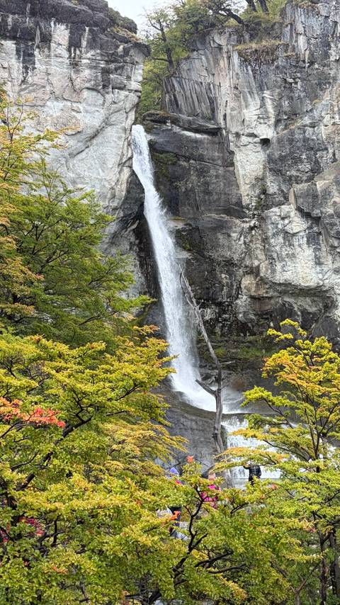       Tall narrow waterfall cascades down a rocky cliff surrounded by early autumn foliage.
  