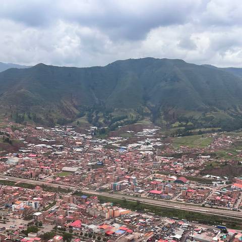       A sprawling Andean town of red roofs fills a valley below towering green mountains under a cloudy sky.
  