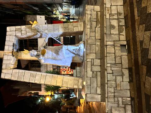       Night-time statue of an Inca ruler holding a staff, illuminated in a small town square.
  