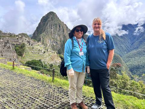      Two travellers smiling with Machu Picchu terraces and lush Andean mountains behind them.
  