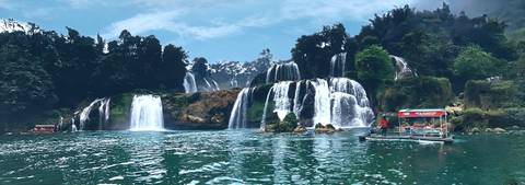       Tiered jungle waterfalls cascade into blue-green pools while visitors approach by small boat.
  
