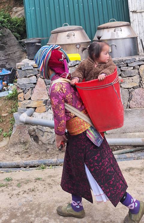       Woman in colourful attire carries a small child in a red basket on her back.
  