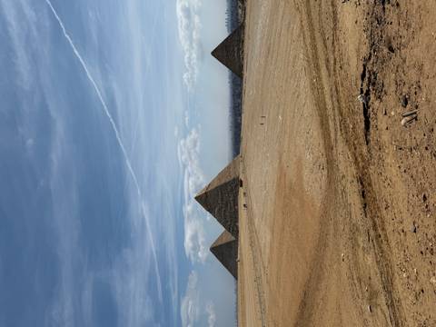       Wide desert vista with the three main pyramids of Giza rising from the sands beneath streaked blue sky.
  