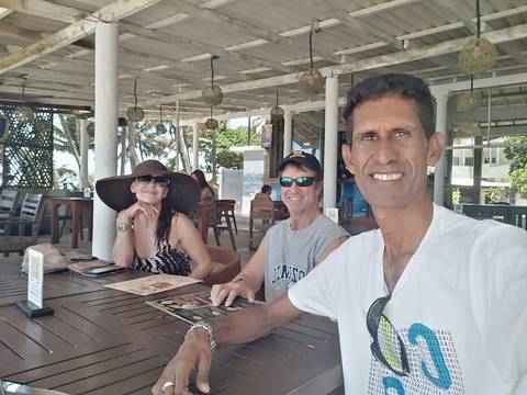       Selfie of guide with two travellers seated at a seaside restaurant under a thatched roof.
  