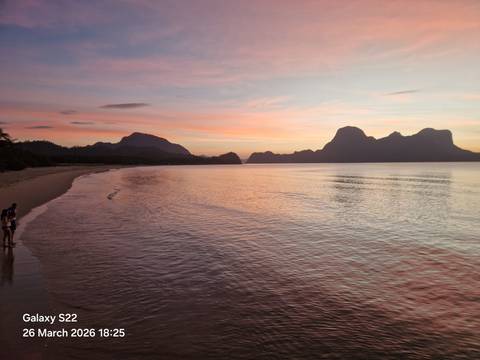       Spectacular pink-orange sunset over a calm tropical bay with karst islands silhouetted on the horizon and two friends standing at the water's edge.
  