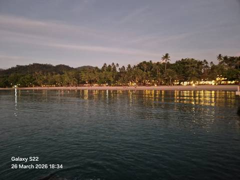       Twilight view from the water of a palm-lined beach illuminated by warm resort lights reflecting on the sea.
  