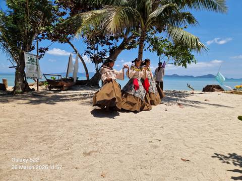       Traditional dancers in flowing brown skirts performing on a sandy beach lined with palms and turquoise water.
  