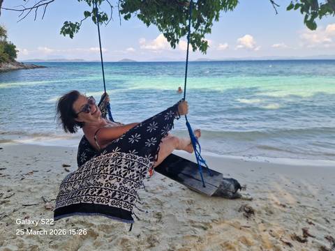       Smiling woman lounging on a rope swing over white sand with clear turquoise sea in the background.
  