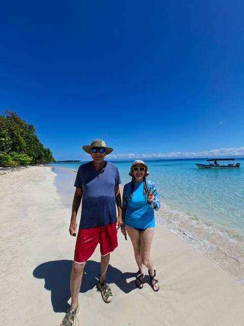       Smiling couple standing on white-sand tropical beach with clear turquoise water and small boat offshore
  