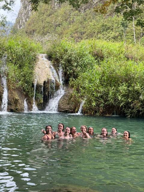       Nine travelers smile while swimming in a turquoise pool beneath a small waterfall and lush greenery.
  