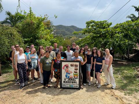       Large tour group poses with a community signboard against a backdrop of green hills and forest.
  