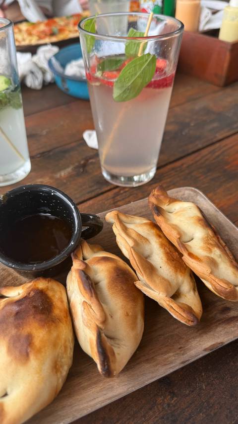       Wooden plate with baked empanada pastries beside a small mug of dark dipping sauce on a rustic table.
  