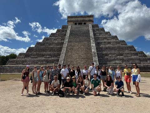       Tour group stands in front of the grand stepped pyramid of Chichen Itza under bright blue skies.
  