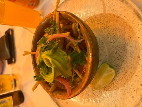       Earthen bowl of hearty soup topped with avocado slices, herbs and pickled onions under warm lighting.
  