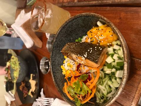       Colorful bowl of salmon, mango, vegetables and sesame seeds alongside a cocktail on a wooden table.
  