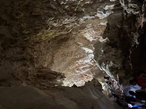       Interior of a large limestone cave with dramatic formations, dim light and visitors exploring a walkway.
  