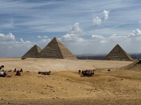       Panoramic view of the three Great Pyramids of Giza rising from the desert plateau under scattered clouds.
  