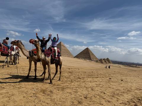       Two travelers on decorated camels raise their arms joyfully with the pyramids behind them.
  
