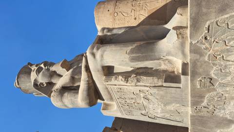       Close-up of a large seated pharaoh statue against a clear blue sky at an ancient Egyptian temple.
  