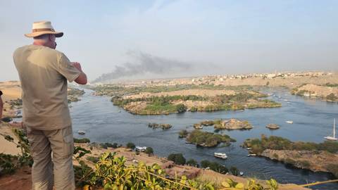       Man in a hat photographing a wide bend of the Nile River with desert hills and city in the distance.
  