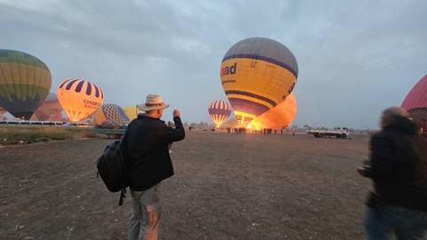       Several colorful hot-air balloons being inflated at dawn while visitors watch on the dusty launch field.
  