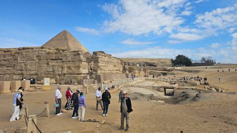       Wide scene of Giza Plateau with pyramid, ruins and scattered tourists under a partly cloudy sky.
  