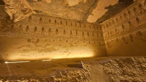       Dimly lit tomb chamber walls covered with detailed ancient Egyptian hieroglyphs and figures.
  