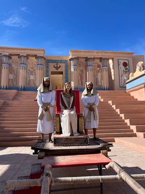       Tourist seated on a throne flanked by two men in ancient Egyptian costumes at a movie set.
  