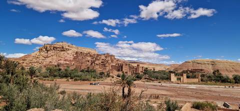       UNESCO mud-brick ksar of Ait Benhaddou set against blue sky and scattered palm trees.
  