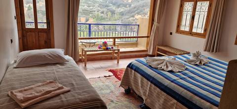       Cozy guesthouse room with twin beds, striped blankets and balcony overlooking green mountain valley.
  