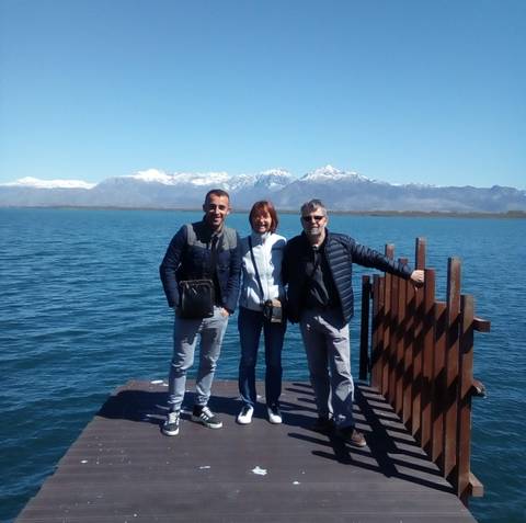       Three friends pose on a wooden pier with a blue lake and snow-capped mountains behind them.
  