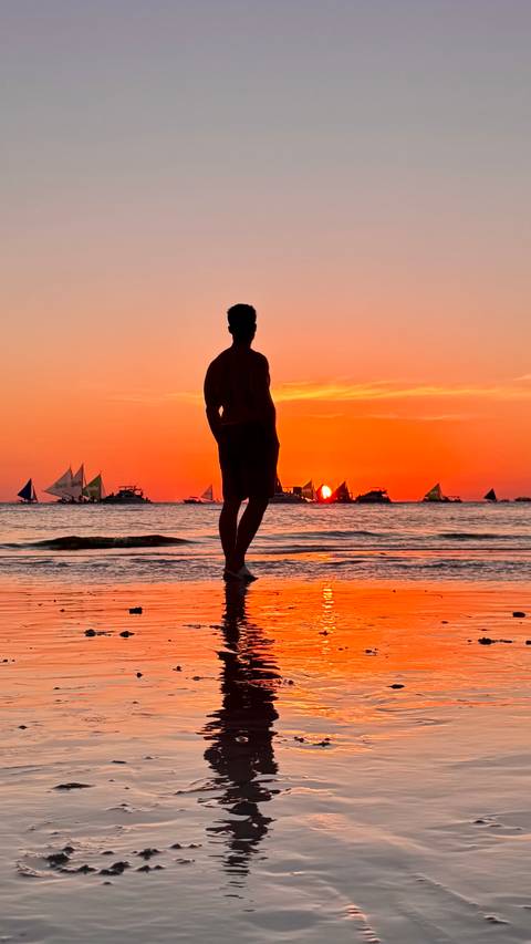       Silhouette of a man on the beach watching a vivid orange Boracay sunset with sailboats.
  