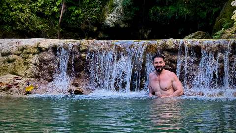       Man relaxes waist-deep in a natural pool beneath a small tropical waterfall surrounded by lush greenery.
  
