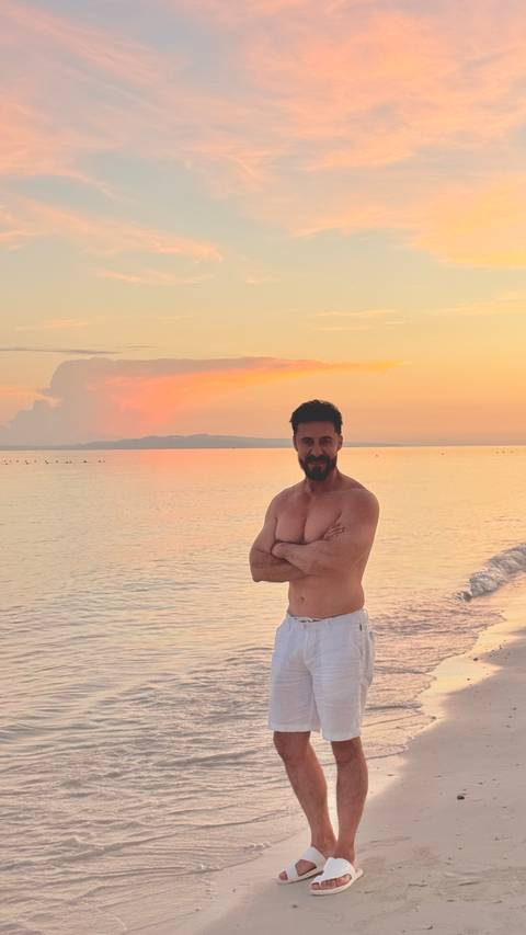      Shirtless man stands on a calm beach at colorful sunset with gentle waves and distant island silhouette.
  